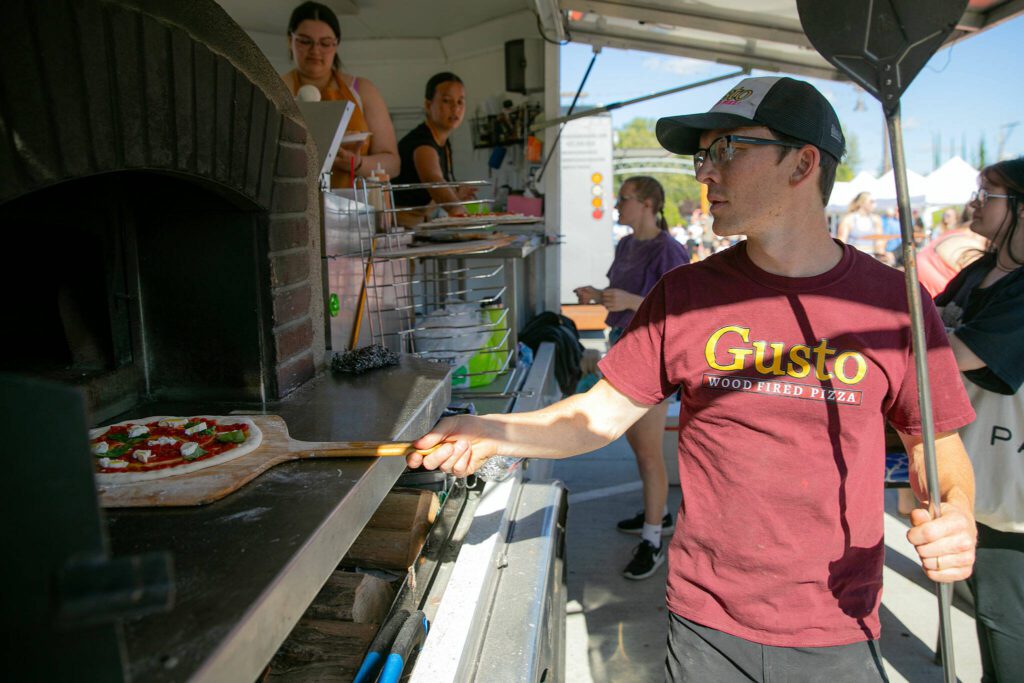 Zach Meyer, who owns Gusto Wood Fired Pizza with his wife Lindsay, throws a Margherita pizza into the oven while cooking at the Lake Stevens Farmers Market on Wednesday, June 12, 2024, in Lake Stevens, Washington. (Ryan Berry / The Herald)
