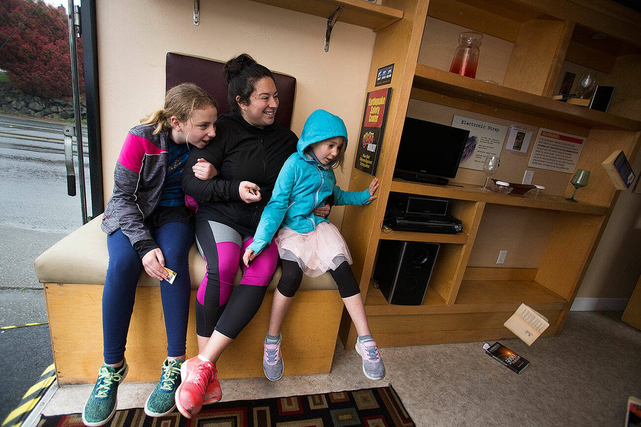 Betty Davis, left, holds on to her mother Leia, who holds onto daughter Alana, 7, as they watch books fall from shelves in the inside The Big Shaker as it shakes like a magnitude seven-plus earthquake on April 19, 2019, in Everett. The Big Shaker is an earthquake simulator in a 24-foot trailer that replicates a typical living room of sofas and shelves with books and plastic dishes that fall during the demonstration. (Andy Bronson / The Herald)