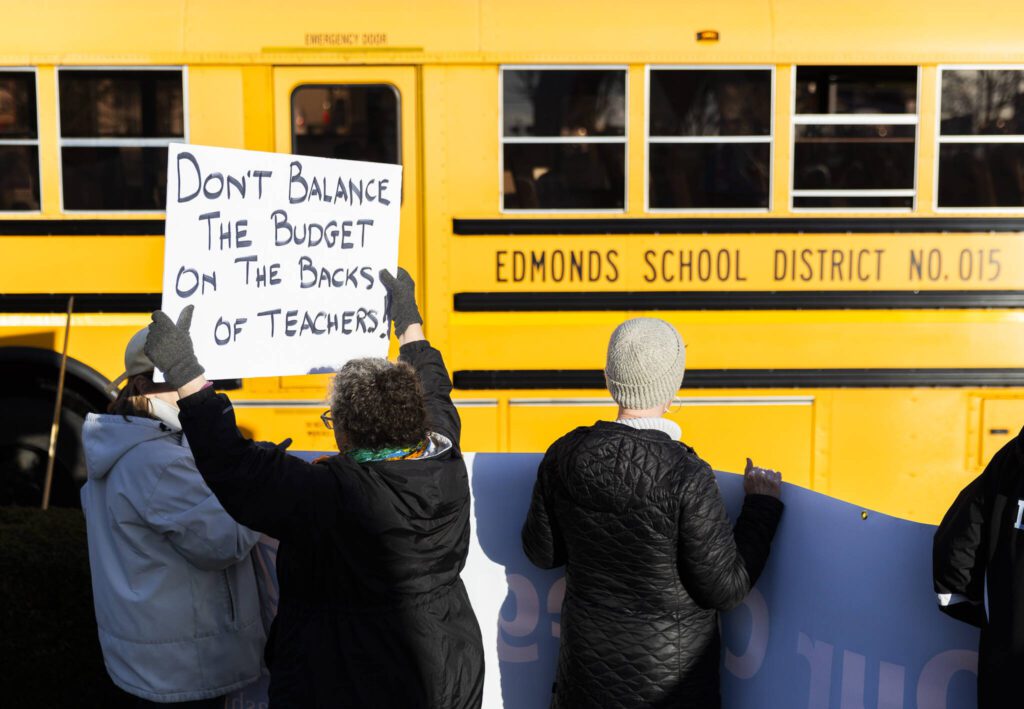 Melody Schneider holds a sign protesting pay cuts to teachers as an Edmonds School District bus passes by during Edmonds College faculty union rally as part of a national day of action outside of the Lynnwood Event Center on Tuesday, March 4, 2025 in Lynnwood, Washington. (Olivia Vanni / The Herald)
