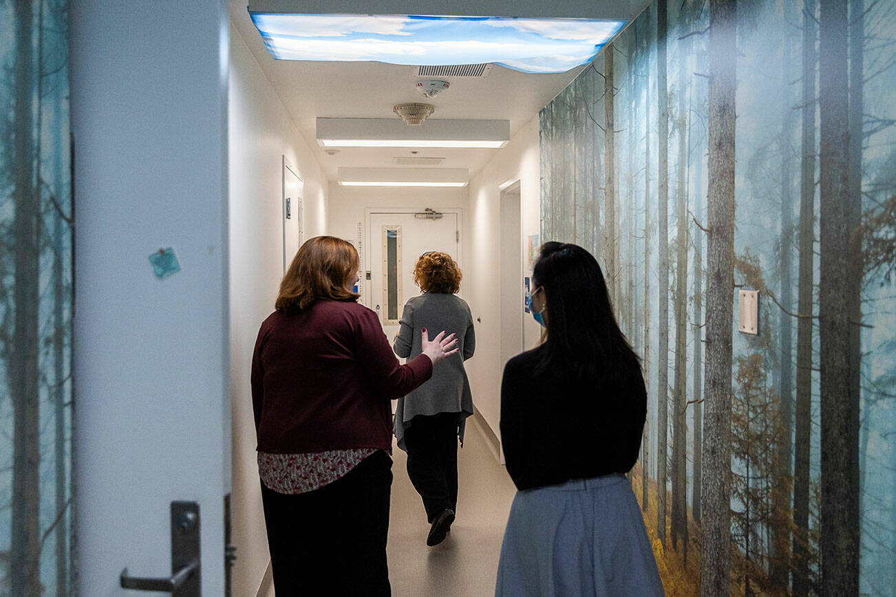 Dr. Katie Gilligan walks down a hallway with forest wallpaper and cloud light shades in the Mukilteo Evaluation and Treatment Center with Amanda Gian, right, and Alison Haddock, left, on Monday, March 24, 2025 in Mukilteo, Washington. (Olivia Vanni / The Herald)