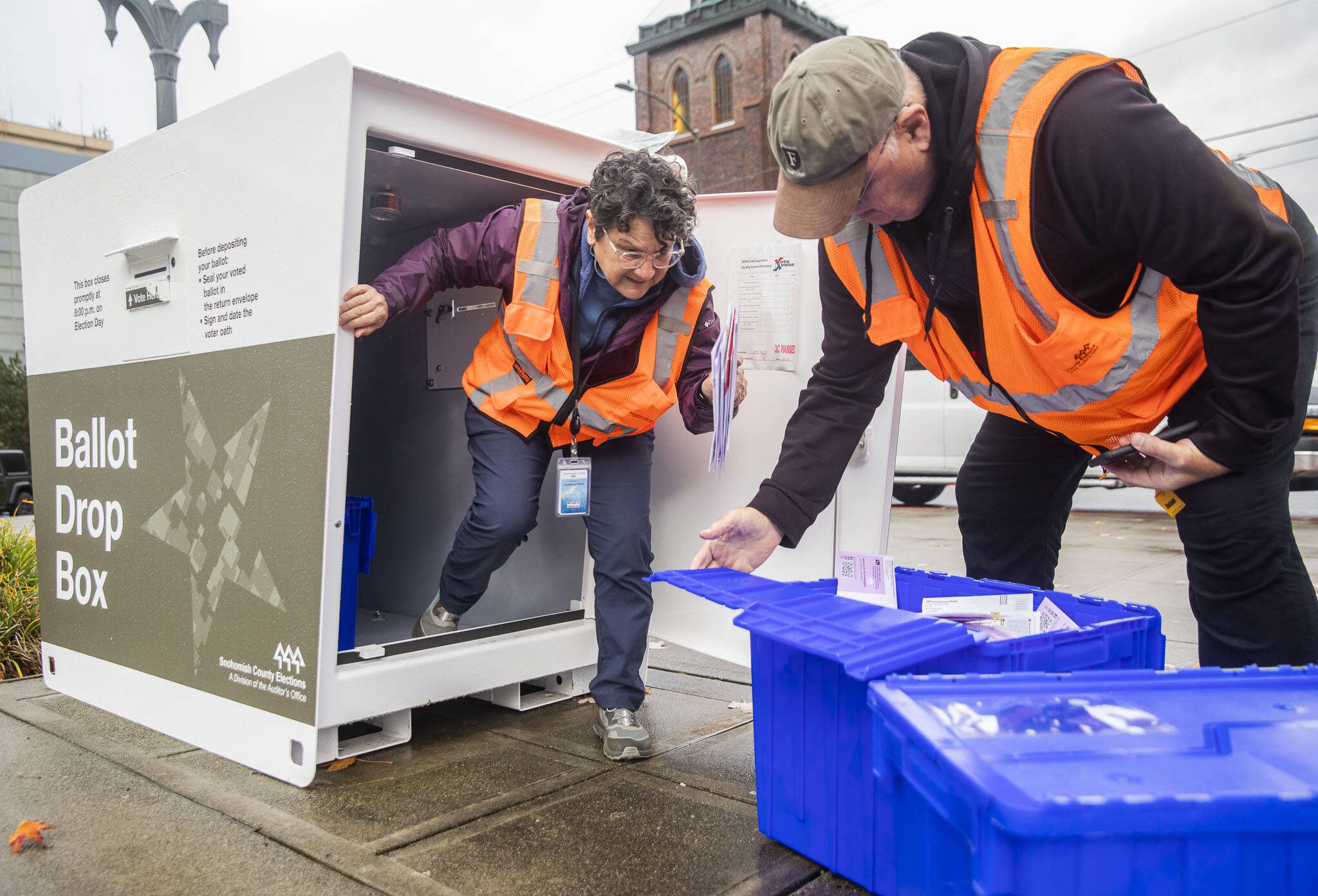 Snohomish County Elections employees Alice Salcido, left and Joseph Rzeckowski, right, pull full bins of ballots from the Snohomish County Campus ballot drop box on Monday, Nov. 4, 2024 in Everett, Washington. (Olivia Vanni / The Herald)