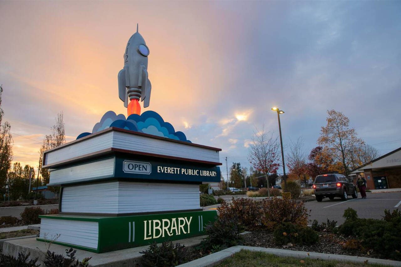 The sun sets beyond public art of stacked books at the Evergreen Branch of the Everett Public Library as a patron returns books in November 2022, in Everett. (Ryan Berry / The Herald file photo)