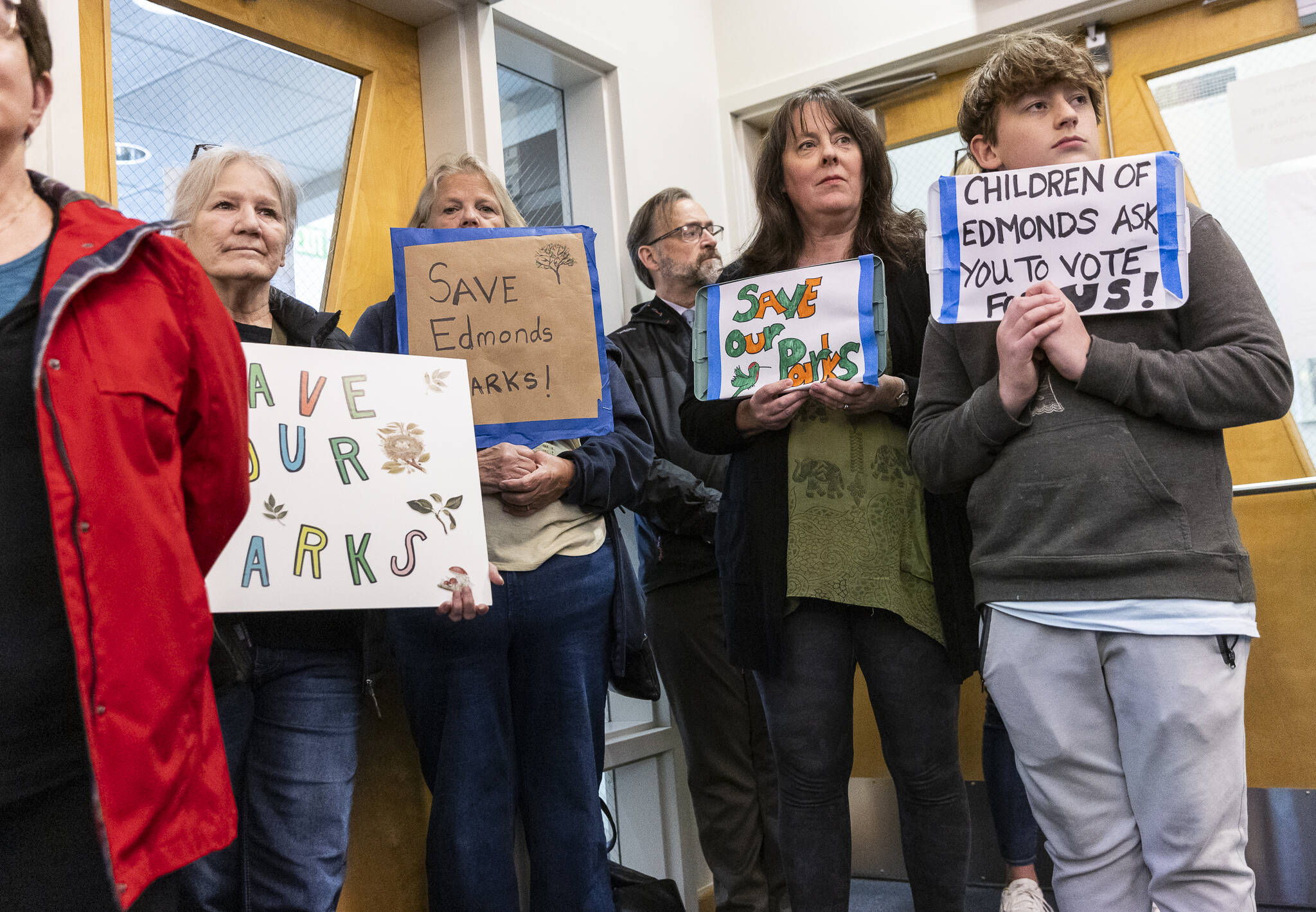 Sue Garton, left to right, Bonny McMahon, Kassie Goforth and Jack Perry stand in the Edmonds City Council chamber holding signs in protest of the idea of selling Hummingbird Hill Park on Monday, April 21, 2025 in Edmonds, Washington. (Olivia Vanni / The Herald)