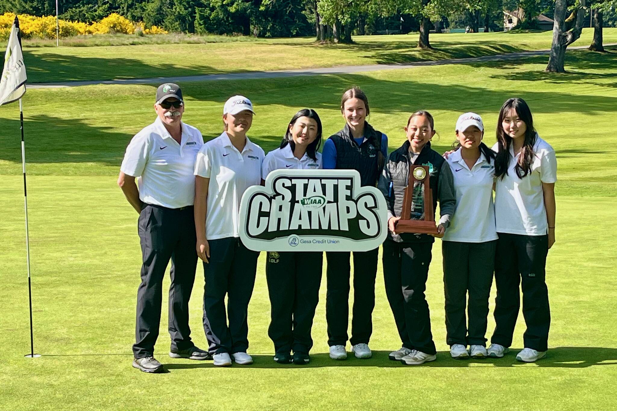 The Jackson girls golf team poses with the state championship trophy and sign on the 18th green of Eagle's Pride Golf Course after winning the WIAA 4A State Championship in DuPont, Washington on May 21, 2025. Pictured left to right: Coach Jerome Gotz, freshman Karen Shin, sophomore Kayla Kim, senior Paige Swander, senior Lindsay Catli, sophomore Chanyoung Park and junior Christine Oh. (Joe Pohoryles / The Herald)
