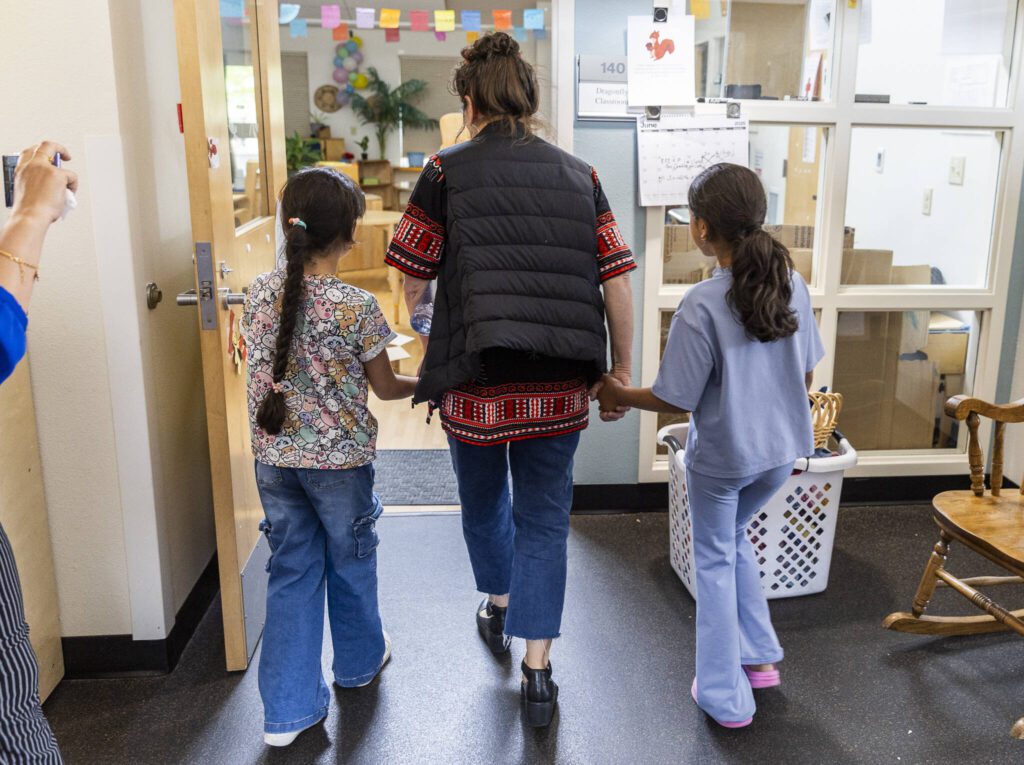 Preschool teacher Michelle Depew walks hand in hand with former Early Learning Center students Lareen Almadjidi, 7, left, and Qamar Almadjidi, 9, right, as they explore their old classroom on Wednesday, June 25, 2025 in Everett, Washington. (Olivia Vanni / The Herald)
