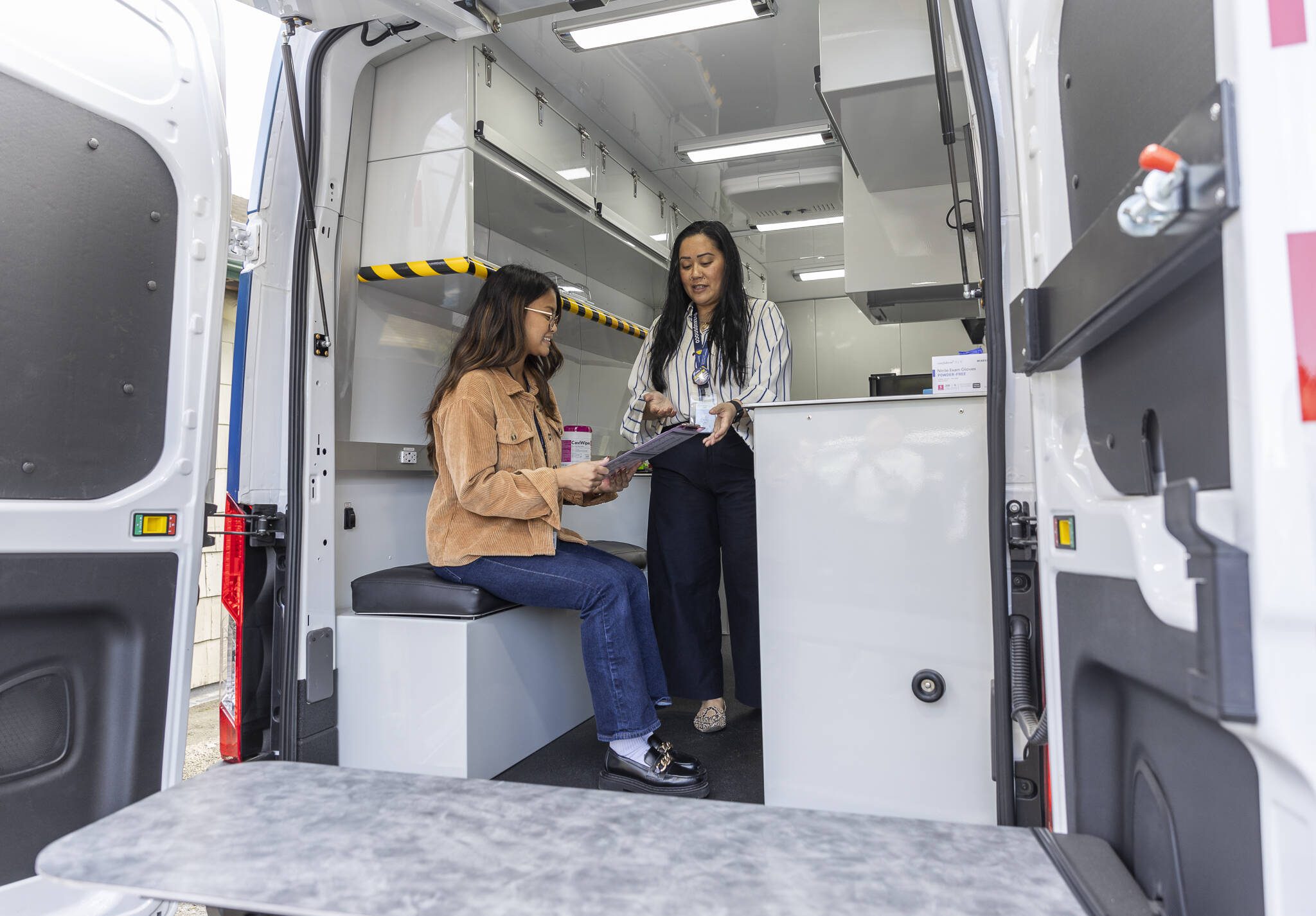 Pia Sampaga-Khim, right, and Jana Rafi, left, demonstrate how a patient check-in might go in the Snohomish County Health Department’s new Health on Wheels Van on April 9. An email from Interim Department Director Pam Aguilar — sent to all Health Department employees Wednesday — announced a temporary pause on hiring. (Olivia Vanni / The Herald)