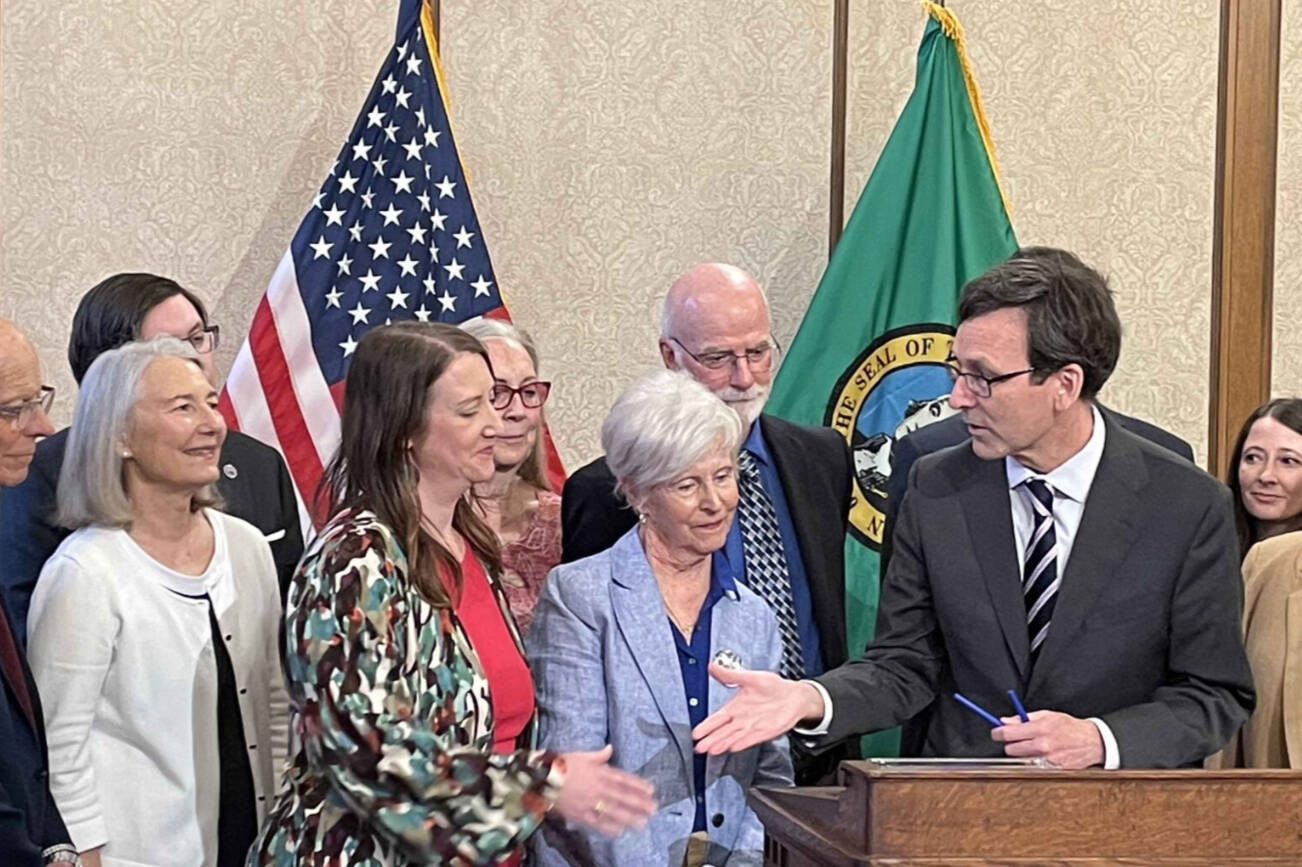 Gov. Bob Ferguson, at podium, goes to shake hands with state Sen. Noel Frame, D-Seattle, at the signing of a bill to make clergy mandatory reporters of child abuse and neglect, on May 2, 2025 in Olympia. Standing between them is Mary Dispenza, a founding member of the Catholic Accountability Project. (Photo by Jerry Cornfield/Washington State Standard)