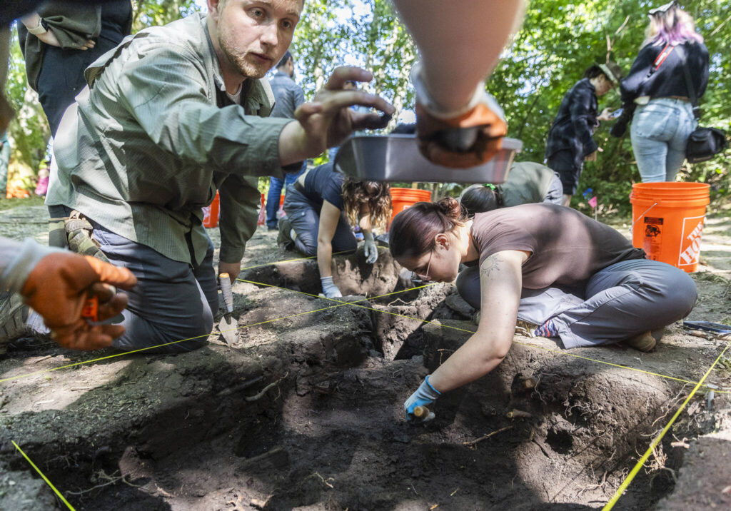 Josh Thiel, left, places a nail into a dust pan while Rey Wall continues digging in a sectioned-off piece of land at Japanese Gulch on Wednesday, July 23, 2025, in Everett, Washington. (Olivia Vanni / The Herald)
