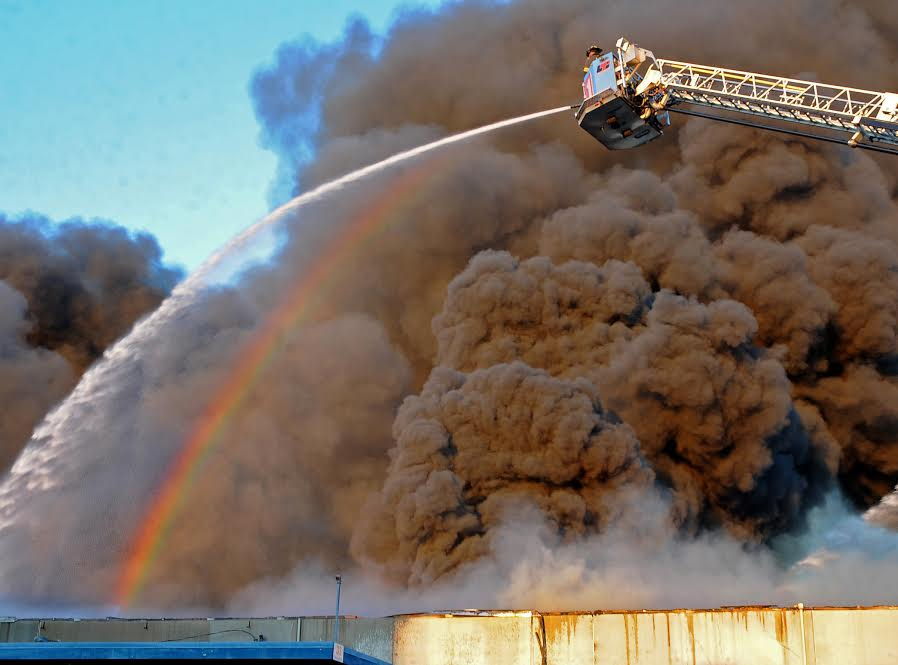 A rainbow emerges within the dark smoke as an Everett firefighter directs a stream of water from atop of Ladder 1. Firefighters from throughout Snohomish County battled a massive three-alarm warehouse fire along the Snohomish River in north Everett on Saturday