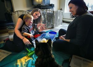Betty Williams (left) and her 19-month-old son, Kayden, play with a makeshift sandbox, which has cornmeal instead of sand. Parent educator Adrienne Matthias (right), supervisor of ChildStrive&rsquo;s Parents as Teachers program, brought the activity to Williams&rsquo; Everett apartment Wednesday as part of her regular home visit. The family cat, Max, kept his distance.