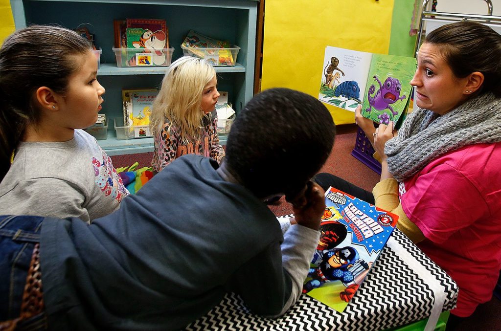While first-graders Carlie Duenas-Jobsky (left) Shelby Spencer and Luhizo Luhizo (foreground) watch and listen, Volunteer Megan hyde (right) reads one of the books. Luhizo Luhizo has his chosen book Captain America to the Rescue in front of him and appears eager to hear it read. (Dan Bates / The Herald)