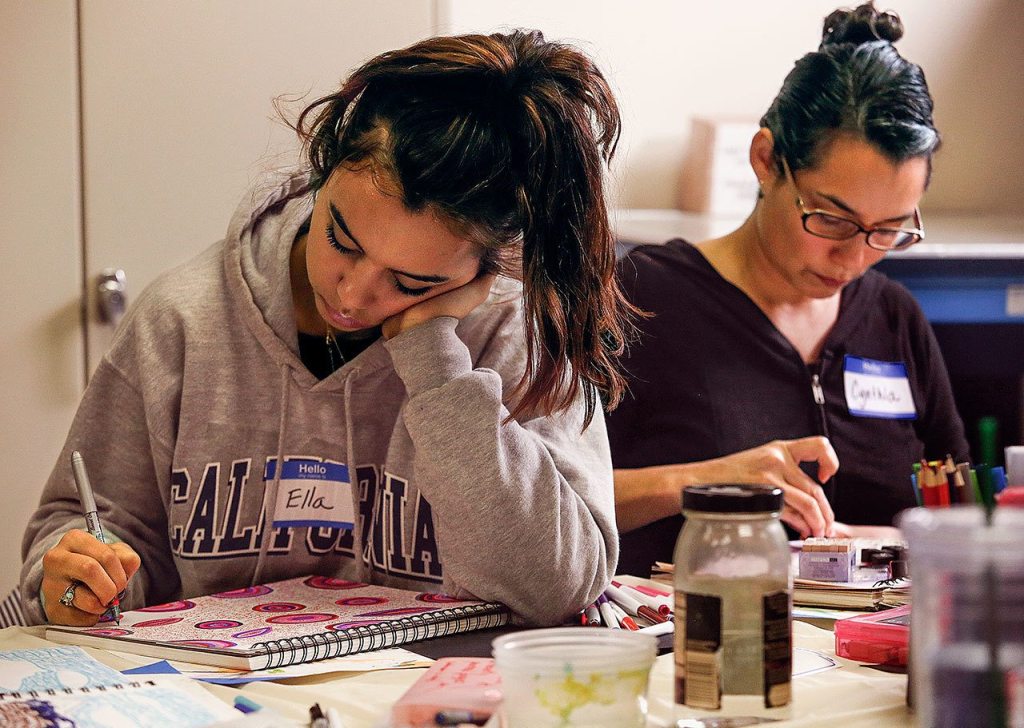 Ella Estlund (left) writes with tiny letters in the spaces around graphic designs she created using a fine-point pen. Cynthia Gahan uses rubber stamps to apply the letters of words in her journal. (Dan Bates / The Herald)
