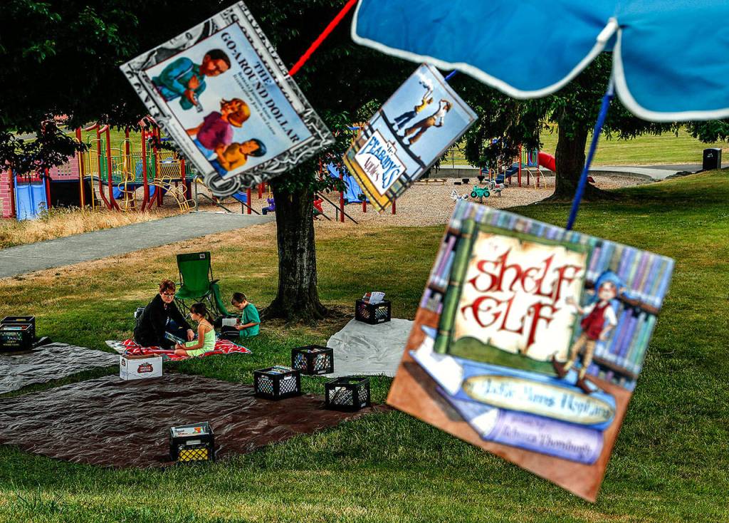Children&rsquo;s book sleeves hang from a sun umbrella and blow in the breeze while Hawthorne Elementary School Assistant Principal Valerie Yob sits on a blanket beneath a tree and talks with Kortana Garner, 8. Kortana&rsquo;s brother, Aaden, 7, reads nearby. (Dan Bates / The Herald)