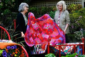 Quilters Kay Davidson (left) and Sydney Hoard test place some of their quilts on an iron bed frame in Janice Tallman&rsquo;s featured garden. The quilt they are lifting up is Hoard&rsquo;s and is titled &ldquo;Dahlias, and Then Some.&rdquo; (Dan Bates / The Herald)