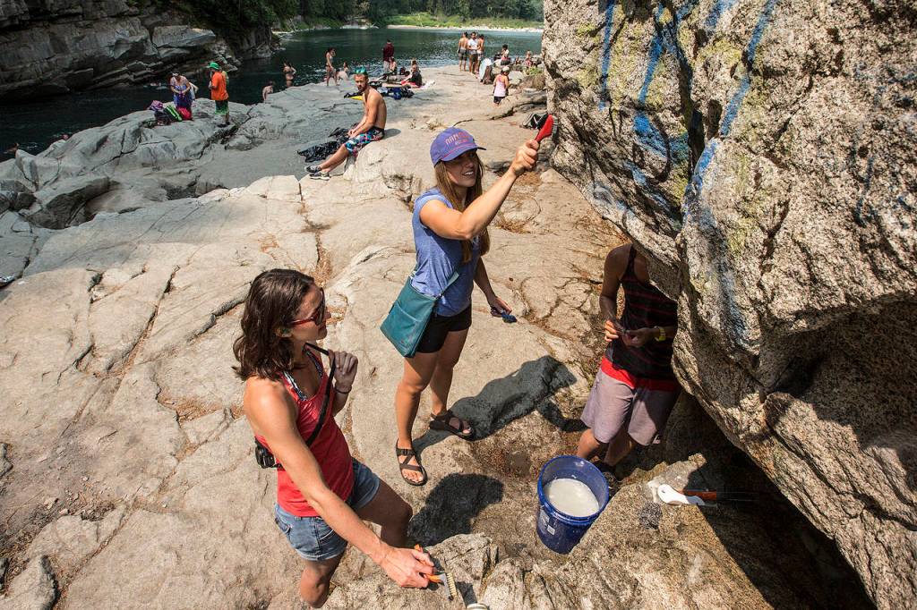 Ally Hinds (center) scrubs away graffiti along with friends Katie Heard (left) and Janelle Dean at Eagle Falls near Index on Saturday, Aug. 5. (Ian Terry / The Herald)