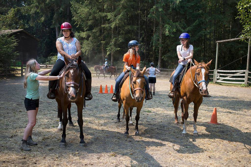 Horse riding staff member Bella Harris (left) talks with Michou Mayer during a riding class at Camp Killoqua on Aug. 2 in Stanwood. McKenzie Levi and Sammi Sutton (right) wait their turn around the arena. (Andy Bronson / The Herald)