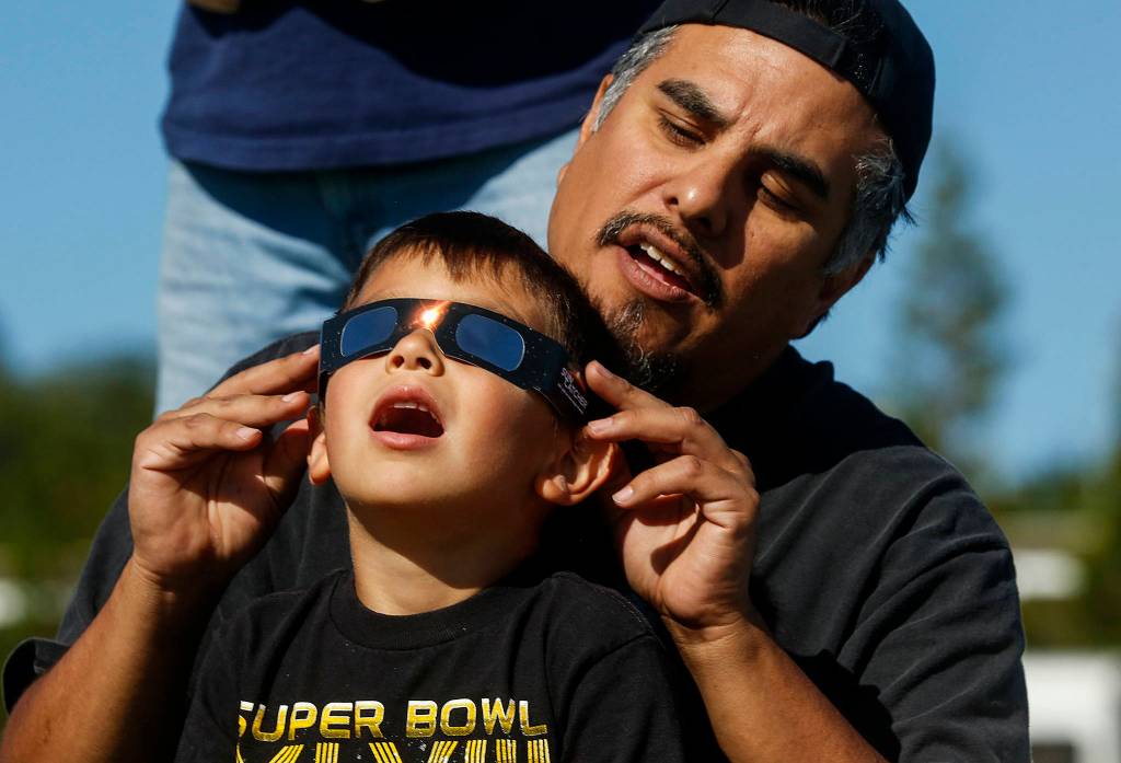 Just off Paine Field near the Future of Flight Aviation Center, Ben Flores of Mukilteo helps his son Vincent, 5, view the solar eclipse through certified safety glasses on Aug. 21. (Dan Bates / The Herald)