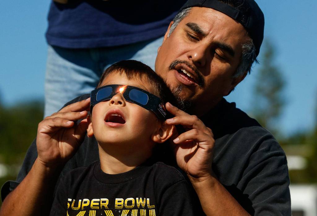 Just off Paine Field near the Future of Flight Aviation Center, Ben Flores of Mukilteo helps his sons, Vincent, 5, and Benny (back), 9, view the solar eclipse through certified safety glasses on Aug. 21. (Dan Bates / The Herald)