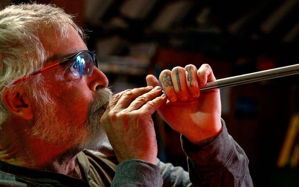Artist Mark Ellinger inflates molten glass with a blowpipe. (Dan Bates / The Herald)