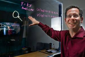 In a lightboard studio, WSU Everett professor Gordon Taub pre-records lectures so he can help students one on one if they need it. He also uses a lightboard in his lectures. It is essentially a large glass surface that he can write and draw on as he speaks face to face with students, as opposed to using a chalkboard with his back to them. It is specifically lit and mounted so content easily stands out against a black background. The video image is flipped horizontally so the symbols and text are not backwords. The system will also allow computer data, photos or pages to share space on the screen with hand-written or drawn content. In the image shown, professor Taub can see the large display screen as it records his teaching efforts.