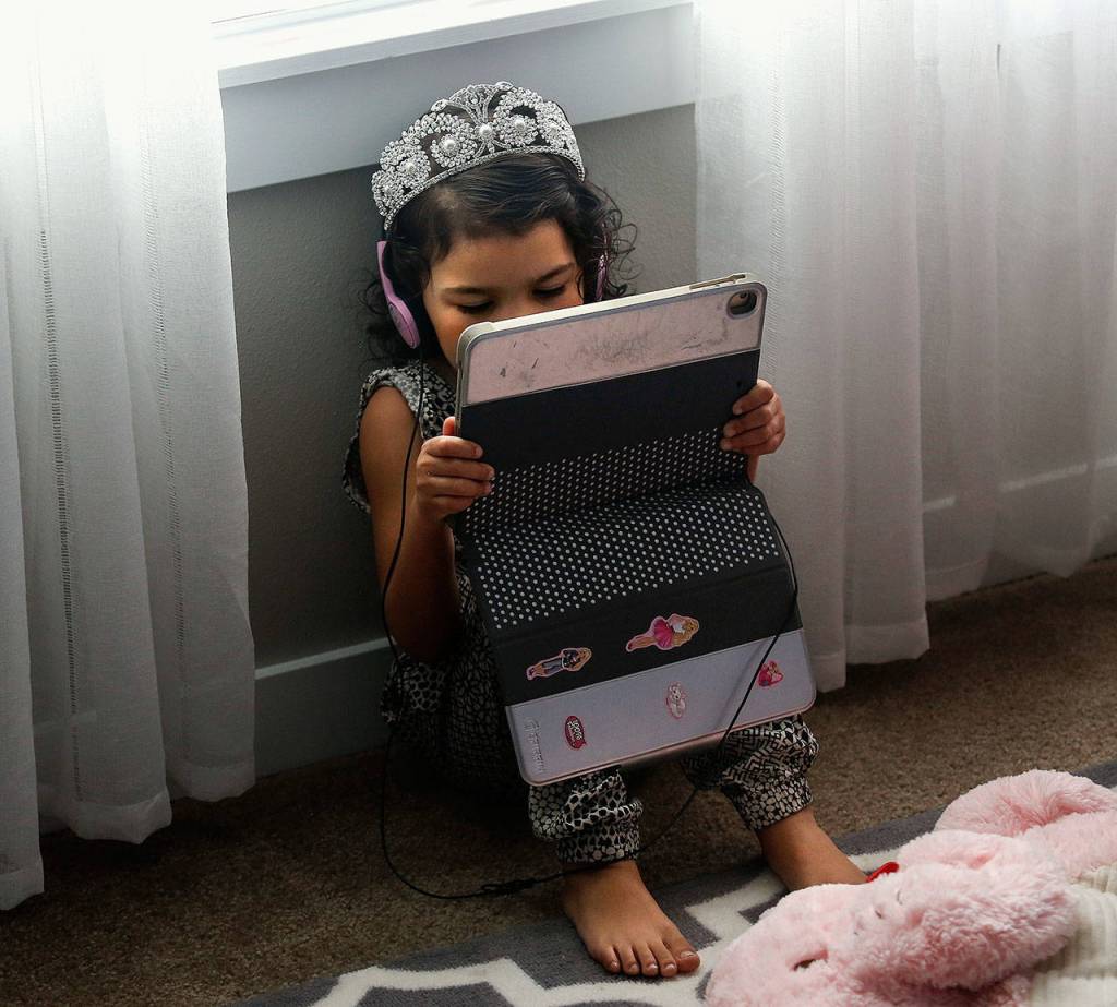 Tiny princess Aria Hansen, 4, appears quite comfortable wearing earphones and her tiara as she sits below a window with an electronic tablet. (Dan Bates / The Herald)