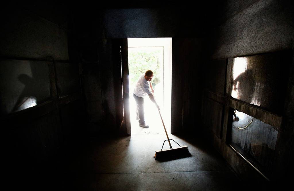 With the heavy door opened and bright morning sunlight splashing in off the granite entry, caretaker Shane Willis sweeps Rucker tomb, likely unaware of his own reflection on glass panels that protect creamation urns along the sides of the tomb. (Dan Bates / The Herald)