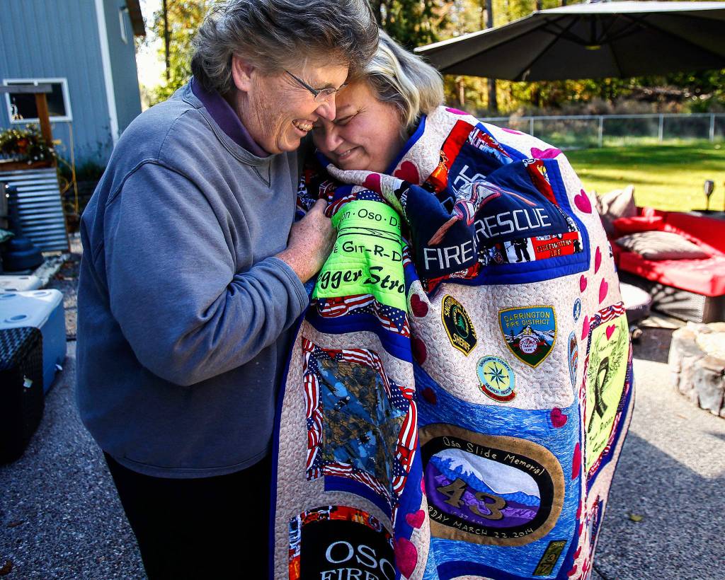 Brenda Willis wraps her friend Karen Pszonka in a quilt that Pszonka commissioned her to create as part of the Oso memorial planning effort. Pszonka lost her daughter, son-in-law and grandchildren in the Oso mudslide. The quilt, made with T-shirts from the agencies involved in the rescue and recovery efforts was the centerpiece of a Sept. 21 <a href="https://www.heraldnet.com/news/oso-memorial-fundraiser-seeks-to-help-build-a-place-to-heal/" target="_blank">fundraiser auction</a>. (Dan Bates / The Herald)