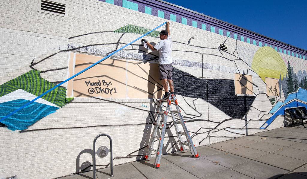 Jake Dkoy Wagoner puts up tape to help him create angles on his spray paint 13-by-75-foot whale mural on a wall of Sound Styles along Main Street on Sept. 28 in Edmonds. (Andy Bronson / The Herald)