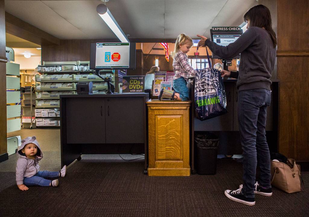 Rachel Hawkins (right) holds open a bag for her daughter Talia Hawkins, 3 (center), to place books in while brother Weston Hawkins, 9 months, sits and waits in Everett Public Library on Nov. 28. (Olivia Vanni / The Herald)