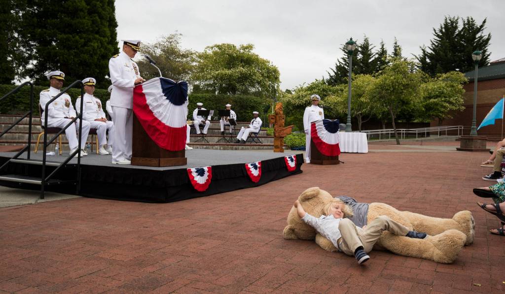 Capt. Mike Davis, the new commanding officer of Naval Station Everett, speaks to the crowd as his son Liam Davis, 3, plays with giant teddy bear during the Change of Command Ceremony on Friday, June 22, in Everett. (Andy Bronson / The Herald)