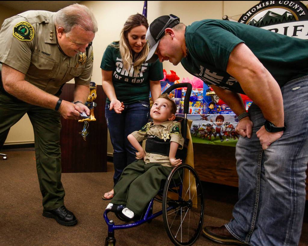From his wheel chair, Isaiah Kobernik, 4, looks up happily as his parents, Gerad and Anastassia Kobernik and Snohomish County Sheriff Ty Trenary lean over him after his dad pinned a shiny new Sheriff badge on his shirt after it was presented to him by Sheriff Trenary at the Snohomish County Courthouse, Tuesday. Isaiah has Spinal Muscular Atrophy, which affects the motor nerve cells in the spinal chord. (Dan Bates / The Herald)