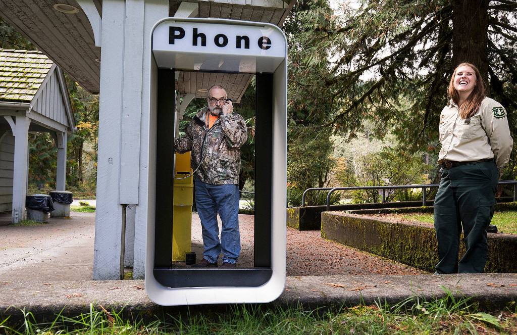 Verlot Corridor Manager Erica Keene laughs as Richard Bake, tech support for Snohomish County, talks to a 911 operator after installing the new pay phone at the Forest Service Building on Oct. 9 in Verlot. The installation by Snohomish County puts a permanent payphone in the town of Verlot, a valuable public safety asset in an area with little cellphone service. (Andy Bronson / The Herald)