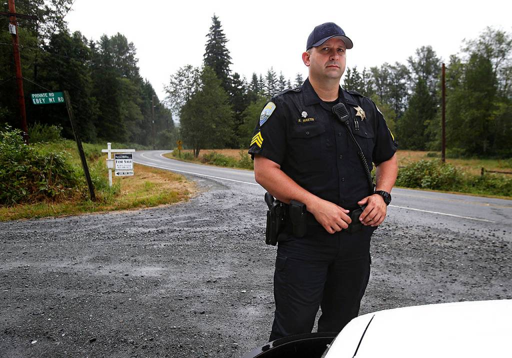 Arlington police Sgt. Rob Martin stands by his car at the intersection of Ebey Mountain Road and Jim Creek Road, where he led a SWAT team to stop Robert Endrizzi, who was shooting at people from behind a berm at the base of the tallest tree (center of picture) in September 2012. (Dan Bates / The Herald)