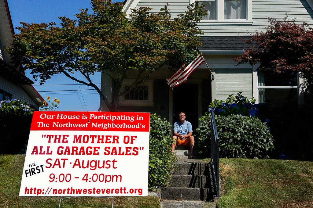 Behind a yard sign in his yard on Rucker Avenue, Bill Loesche takes a break from organizing movies, toys and micro machines he plans to sell Saturday at the Mother of All Garage Sales. (Dan Bates / The Herald)