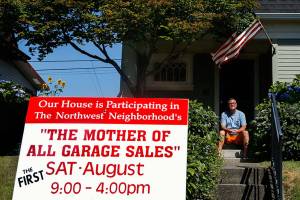 Behind a yard sign in his yard on Rucker Avenue, Bill Loesche takes a break from organizing movies, toys and micro machines he plans to sell Saturday at the Mother of All Garage Sales. (Dan Bates / The Herald)