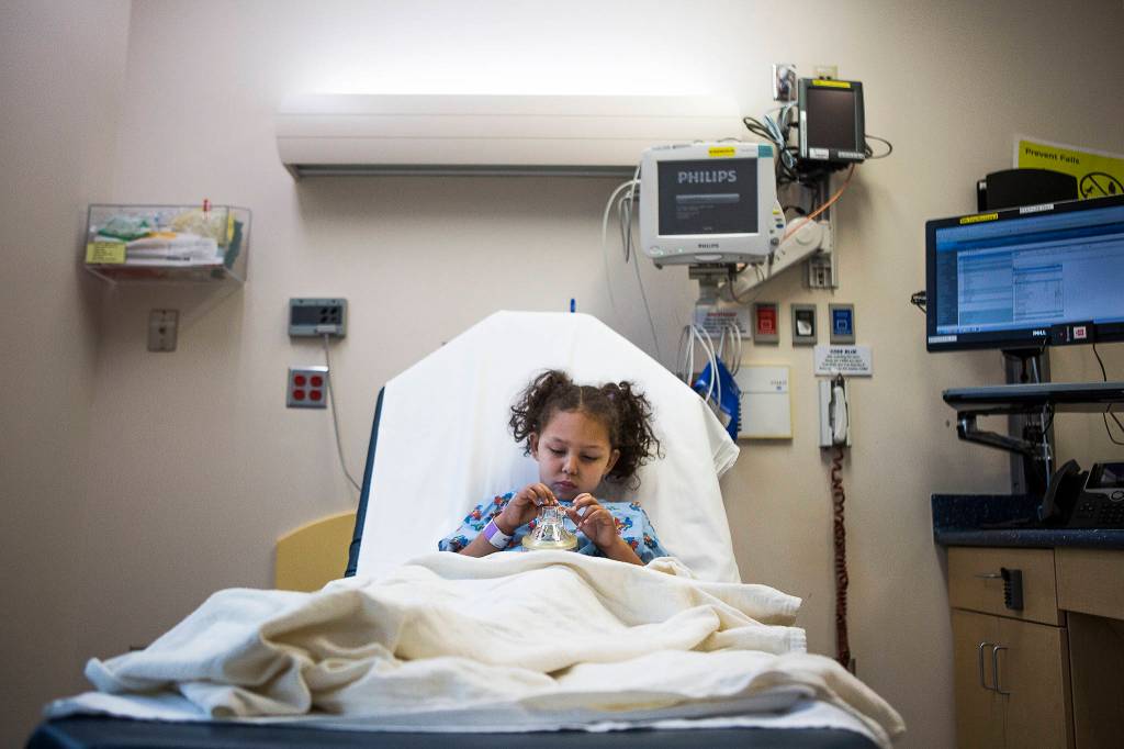 Kaya Sanders, 5, sits in bed at Seattle Childrens Hospital and places car stickers on her anesthesia mask while she waits to be taken in for her MRI on Dec. 12, 2018. (Olivia Vanni / The Herald)