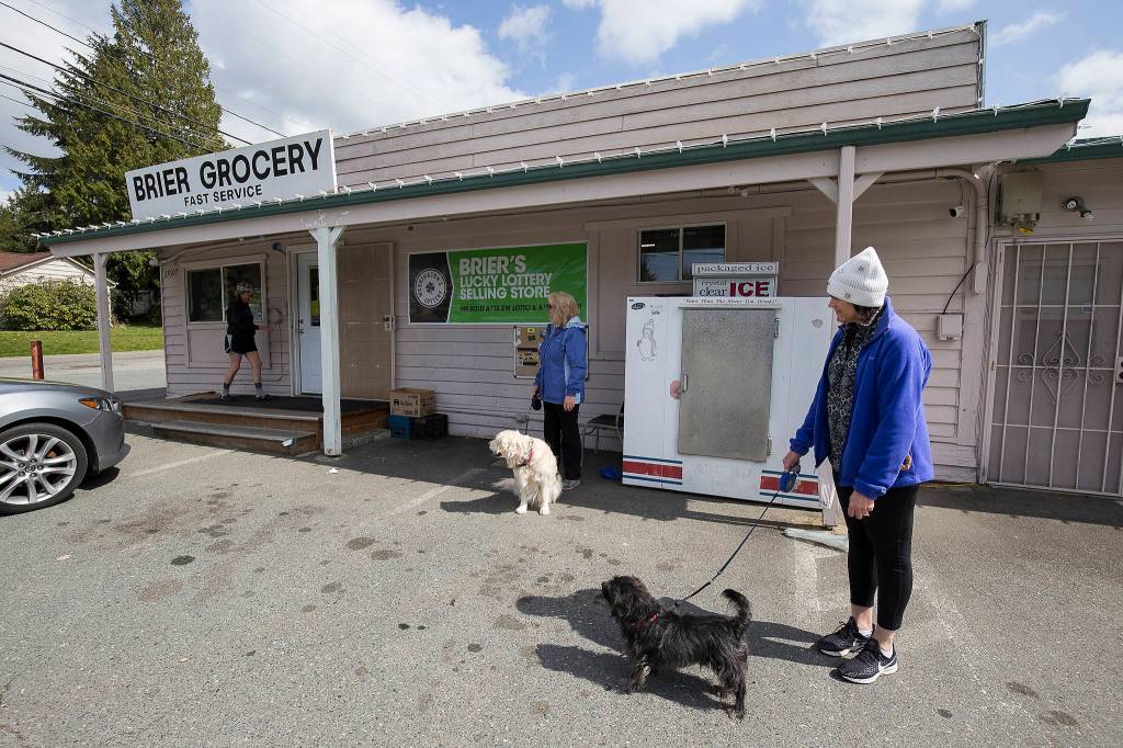 Social distancing themselves from each other, customers Lynne Emmons (center) and Lisa Moen wait for fewer people inside before going into Brier Grocery on April 7 in Brier. (Andy Bronson / The Herald)