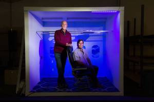 Under UV light, CEO Robert Olson, right, and president Gregory Strand sit in a mockup of an airplane interior on Monday, Sept. 14, 2020 in Everett, Washington. Waypoint Interiors, an Everett aerospace company, is developing an ultraviolet (UV) system to disinfect planes, trains and buses. (Andy Bronson / The Herald)