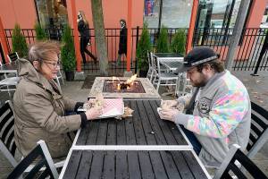 Diners Bonnie Breitman, left, and Casey McGan huddle near an outdoor gas fire as they eat lunch outside in a blustery wind Tuesday, Nov. 17, 2020, in Bellingham, Wash. Washington Gov. Jay Inslee on Sunday announced tighter restrictions in the state in response to a flood of new cases of COVID-19, the disease caused by the coronavirus. Restaurants and bars will again be limited to outdoor dining and to-go service, gyms, and some entertainment centers will be required to close indoor services. Retail stores, including grocery stores, will be ordered to limit indoor capacity and indoor social gatherings will be prohibited unless attendees have quarantined for 14 days or tested negative for COVID-19 and quarantined for a week. (AP Photo/Elaine Thompson)