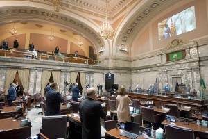 Senate members stand as a ceremonial presentation of colors is done virtually on a video screen above Monday, Jan. 11, 2021, at the Capitol in Olympia, Wash. Washington state's Legislature convened Monday under a large security presence because of concerns about efforts by armed groups who might try to disrupt the proceedings or occupy the Capitol, which is closed to the public due to the ongoing pandemic. (AP Photo/Ted S. Warren)
