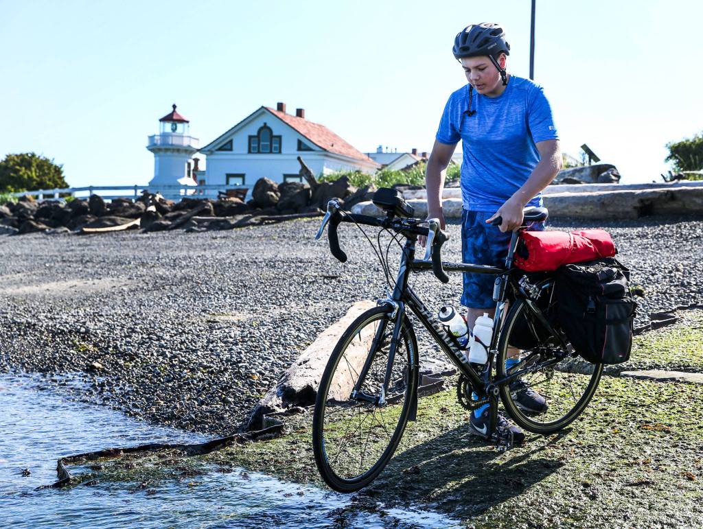 Deven Boyce dips his tire in the water of Puget Sound to start his cross-country cycling trek at the Mukilteo Lighthouse. (Kevin Clark / The Herald)