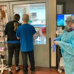 In this Aug. 20 photo, two visitors peer into the room of a COVID-19 patient in the intensive care unit at Salem Hospital in Salem, Oregon, as a nurse dons full protective gear before going into the room of another patient. (AP Photo/Andrew Selsky, File)
