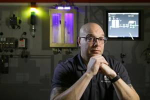 Everett Community College's Dennis Skarr sits in front of a 15-foot interactive wall that can replicate a manufacturing company's assembly line, hardware, software and networks on Wednesday, Nov. 24, 2021 in Everett, Washington. A class taught by Skarr focuses on cyber threats against manufacturers, pipelines, water treatment systems and electrical grids.(Andy Bronson / The Herald)