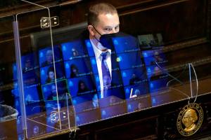 Gary Holt, who reads bills being considered in the Washington House, wears a mask as he sits behind a plexiglass shield with reflections of state representatives meeting remotely on it, Wednesday, April 21, 2021, at the Capitol in Olympia Wash. The House was considering a proposed new tax in Washington state on capital gains that would be imposed on the sale of stocks and bonds in excess of $250,000. (AP Photo/Ted S. Warren)