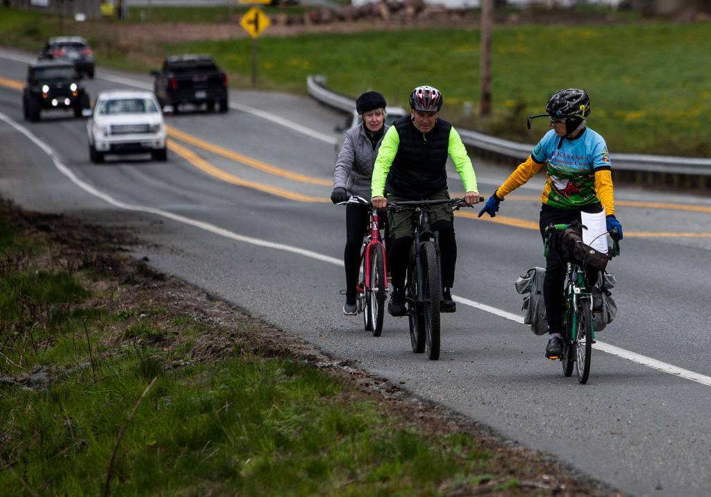 Kristin Kinnamon points to a spot that needs a Dan Henry directional mark as Steve Boskovich and Frances Philbin ride behind along Highway 532 on Sunday, April 10, 2022 in Camano Island, Washington. (Olivia Vanni / The Herald)