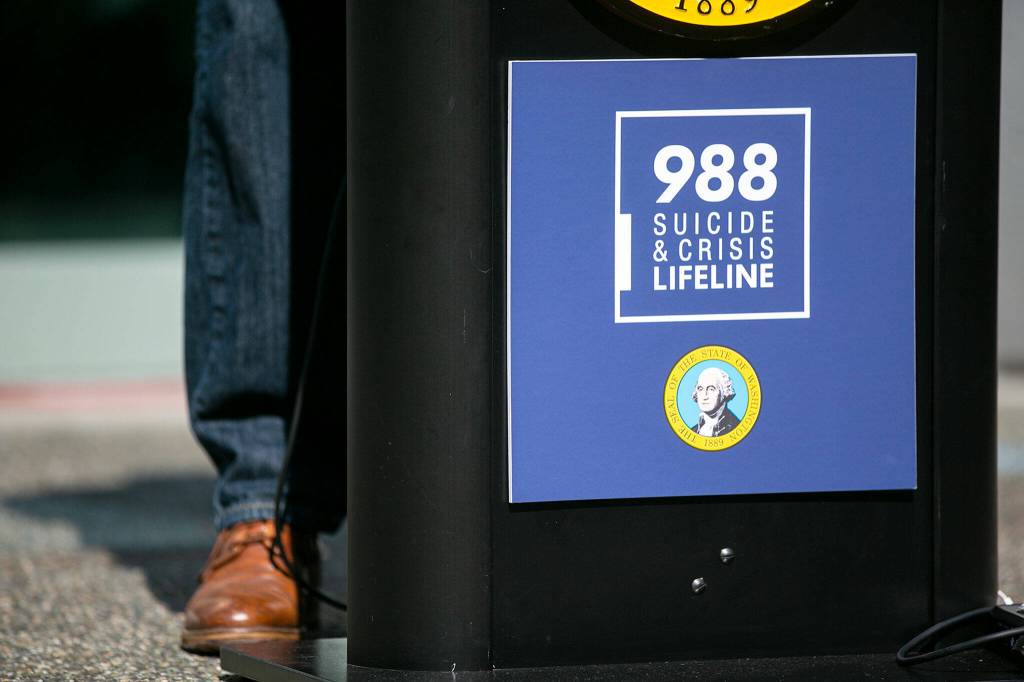 A 988 Suicide & Crisis Lifeline poster hangs from a lectern as Gov. Jay Inslee addresses a crowd about mental health services in Washington on Thursday, in Everett. (Ryan Berry / The Herald)