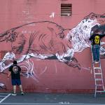 Two artists work on a huge dinosaur mural covering the entire north side of Brews Almighty on Friday, in downtown Everett. (Ryan Berry / The Herald)