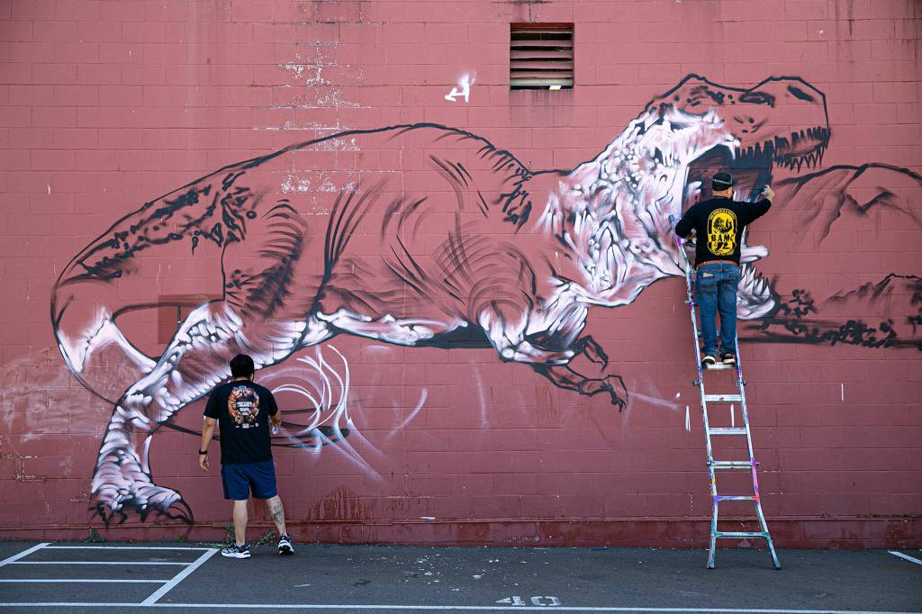 Two artists work on a huge dinosaur mural covering the entire north side of Brews Almighty on Friday, in downtown Everett. (Ryan Berry / The Herald)