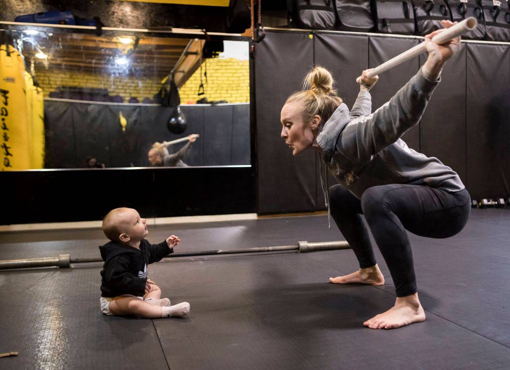 Miranda Granger makes a face at her daughter, Austin, while she does an overhead squat during a warmup in February 2022 at Charlies Combat Club in Everett, Washington. Six months after giving birth, Granger finally ramped up the intensity of her training. In the meantime, she had struggled with fatigue and pelvic floor issues. (Olivia Vanni / The Herald)