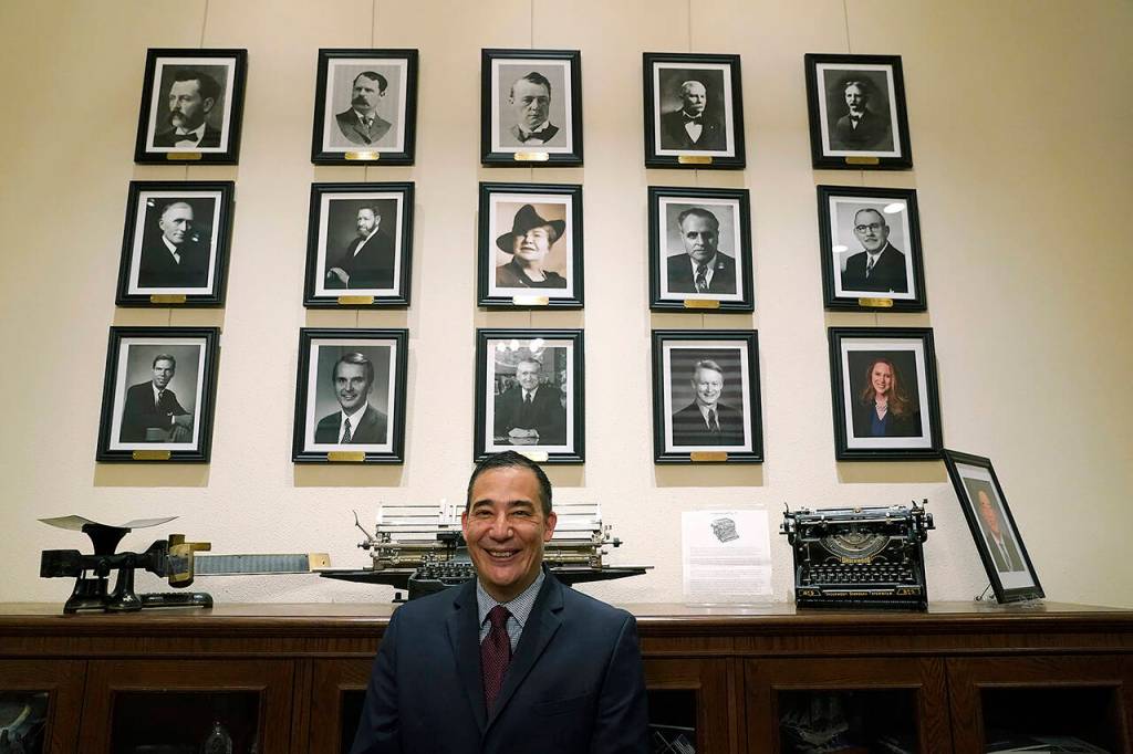 Steve Hobbs, who was sworn in as Washington Secretary of State, Monday, Nov. 22, 2021, at the Capitol in Olympia, poses in front of photos of the 15 people who previously held his office. (AP Photo/Ted S. Warren)
